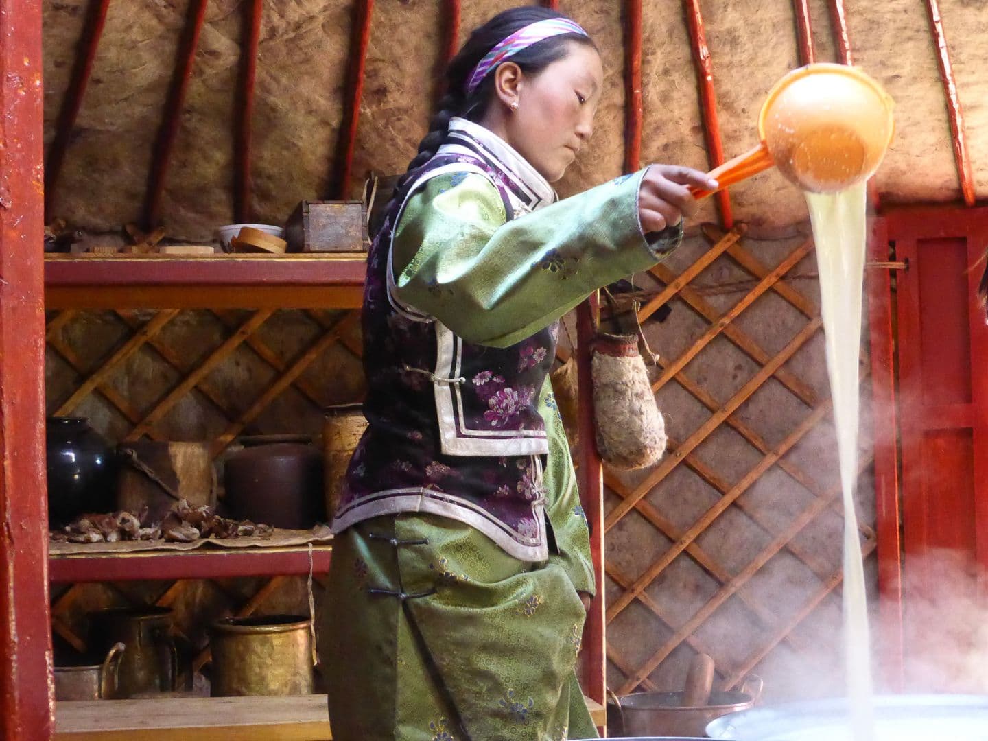 Mongolian woman in traditional clothing pours hot milk tea in a ger, with wooden lattice walls and cooking pots in the background.