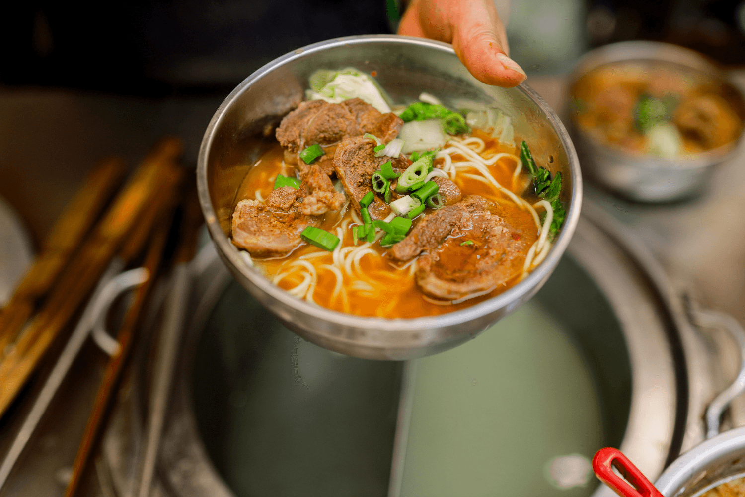 Bowl of Taiwanese beef noodle soup with braised beef, wheat noodles, green onions, and leafy greens in a rich reddish broth.