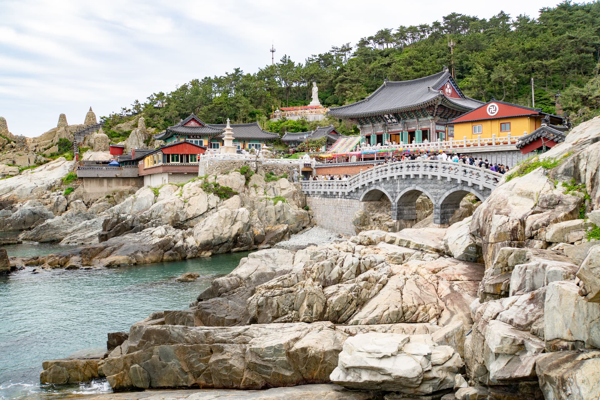 Haedong Yonggungsa Temple on the rocky coastline near Busan, South Korea, with a stone bridge, colorful temple buildings, and visitors overlooking the sea.