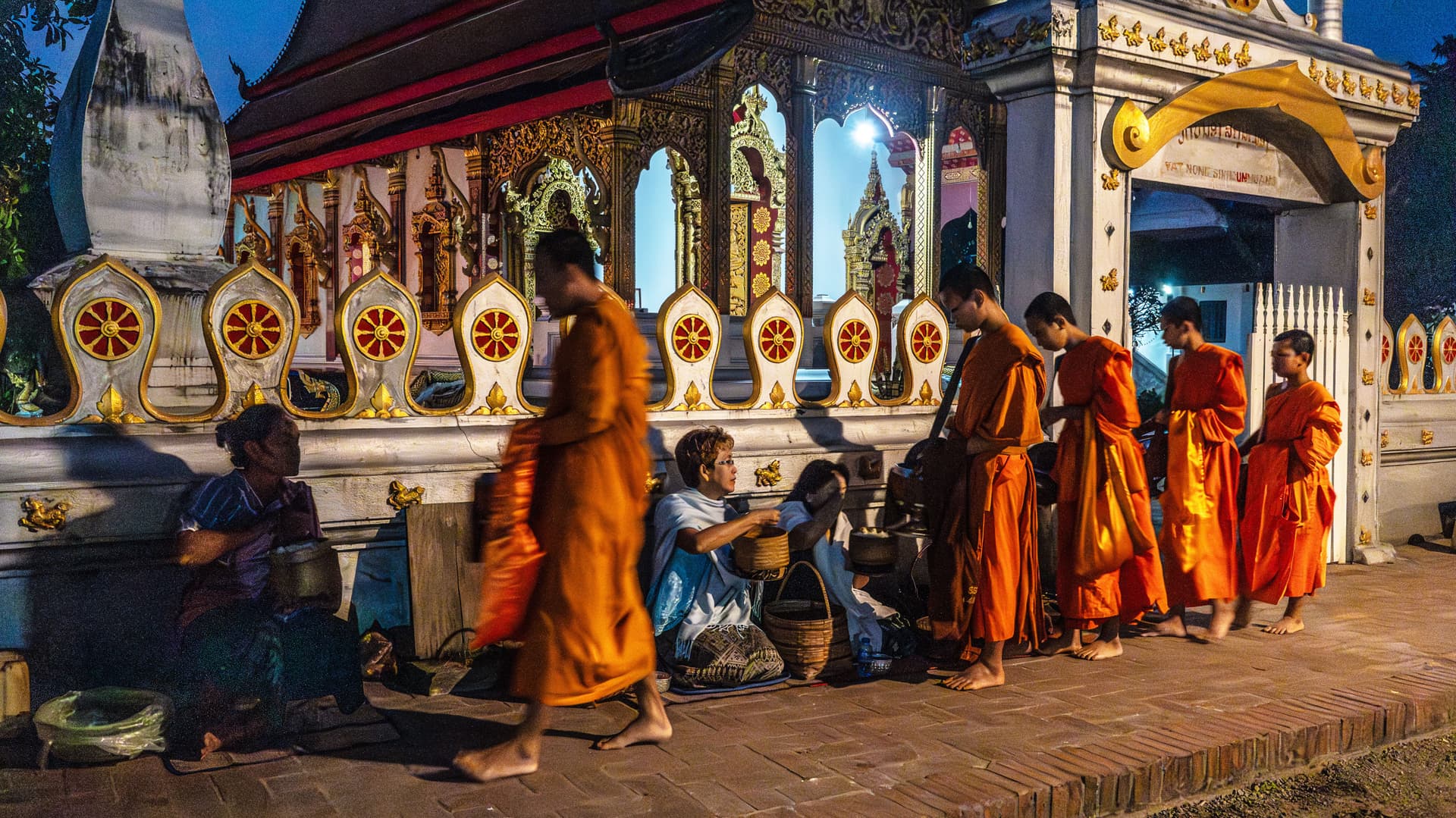 Buddhist monks in saffron robes receiving morning alms from locals outside a temple in Luang Prabang, Laos.