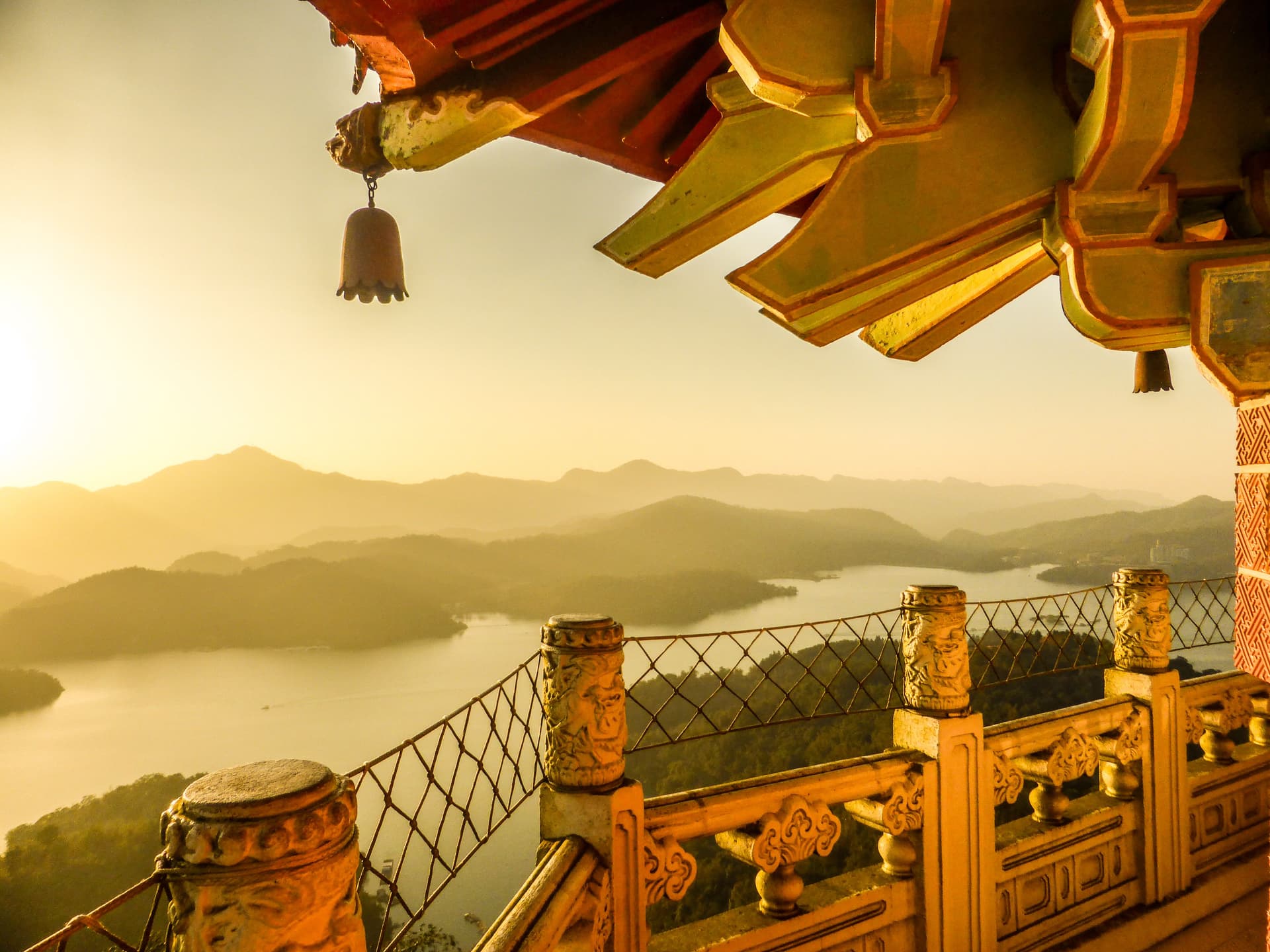Sunrise view over Sun Moon Lake in Taiwan from a temple balcony, with carved stone railing, hanging bell, and misty mountains in the distance.