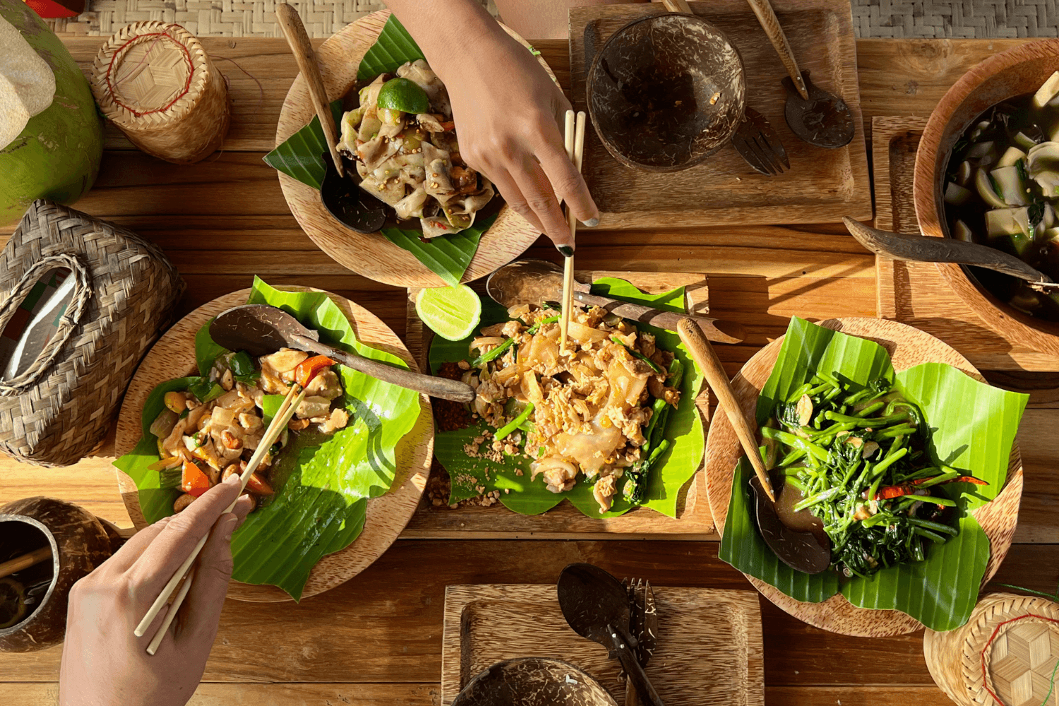 Overhead view of Lao food served on banana leaves on a wooden table, with people sharing dishes using chopsticks, lime wedges, and coconut bowls.