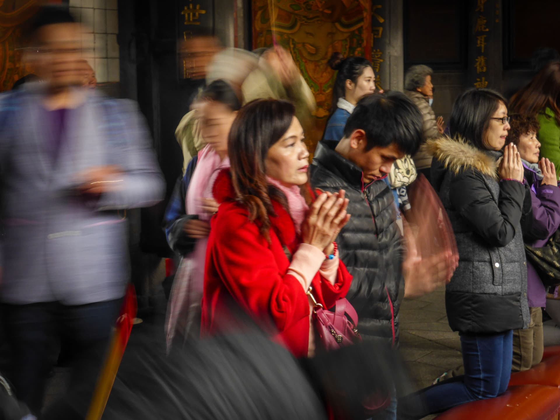 Woman praying with hands clasped inside a Taiwanese temple, with other worshippers around her and motion blur from people passing by.