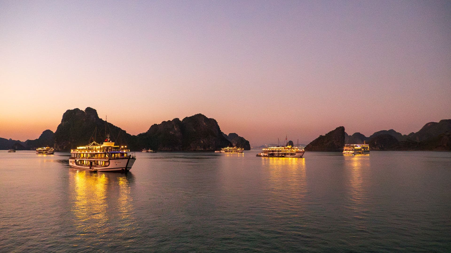 Cruise boats lit up at dusk on calm water in Ha Long Bay, Vietnam, with limestone karst islands silhouetted against a pink sunset sky.