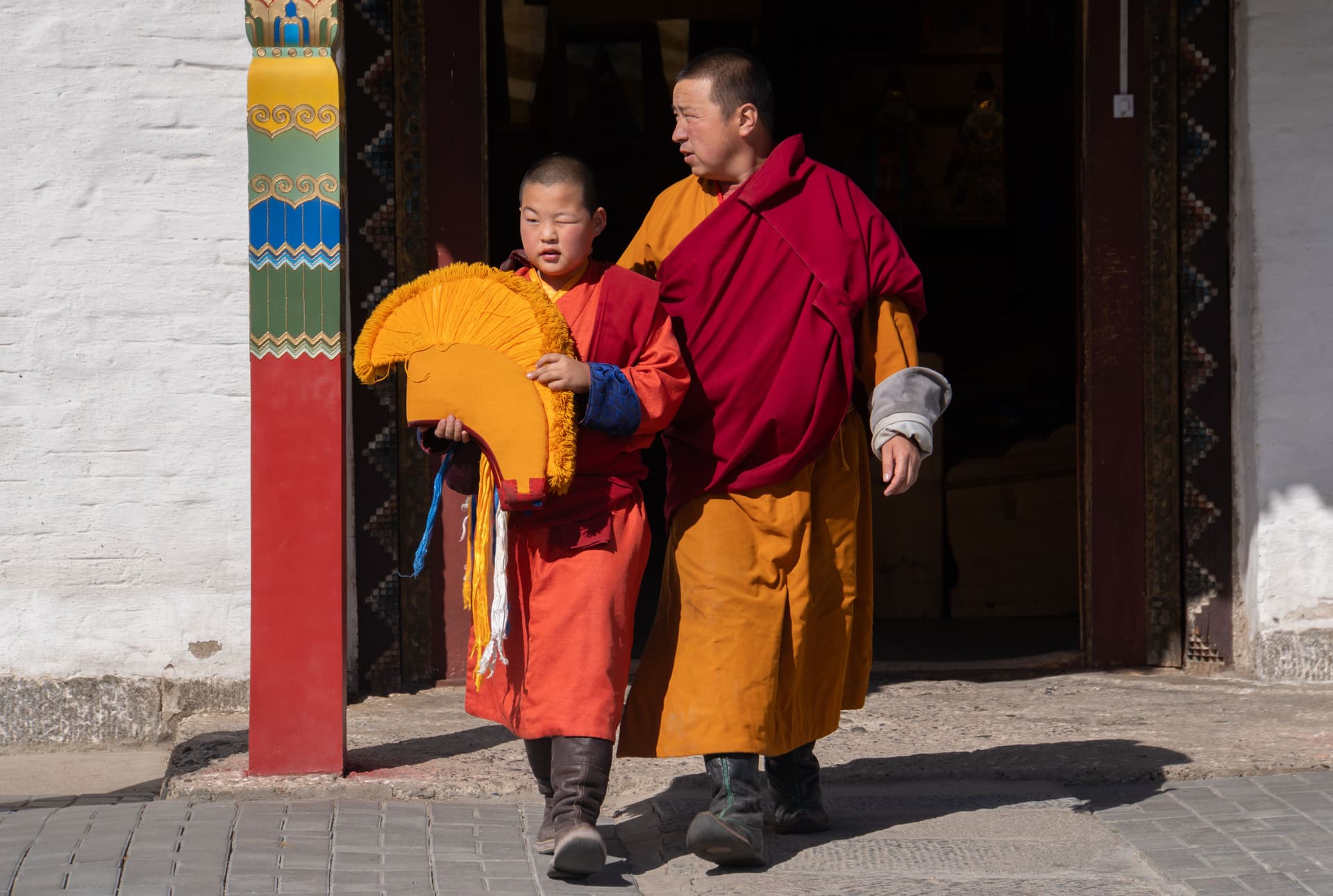 Mongolian Buddhist monk and young novice in red and saffron robes walk out of a monastery doorway, carrying a traditional ceremonial hat.