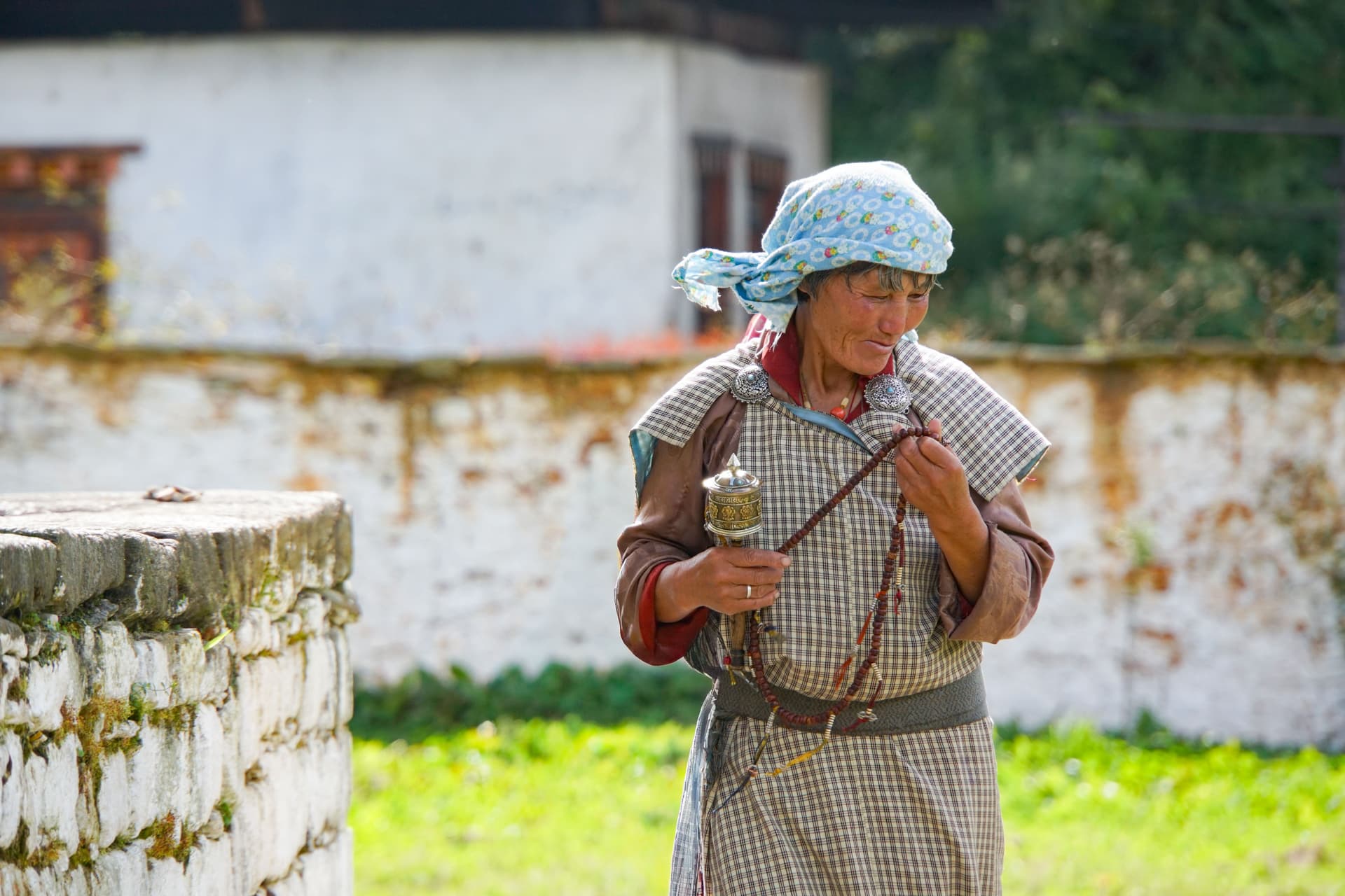 Bhutanese woman in traditional dress and blue headscarf walks in a monastery courtyard holding prayer beads and a handheld prayer wheel.