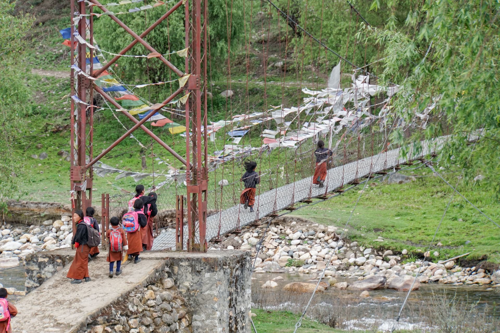 Bhutanese schoolchildren in traditional dress cross a suspension bridge lined with prayer flags over a rocky river in a green valley.